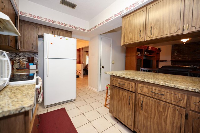 kitchen featuring brown cabinets, light tile patterned floors, backsplash, freestanding refrigerator, and under cabinet range hood