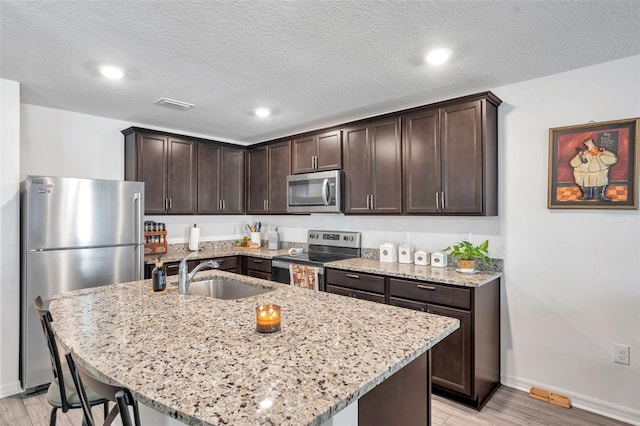 kitchen featuring a kitchen island with sink, sink, dark brown cabinets, light stone counters, and appliances with stainless steel finishes