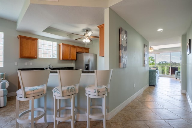 kitchen featuring light tile patterned flooring, a ceiling fan, stainless steel refrigerator with ice dispenser, brown cabinetry, and a kitchen bar