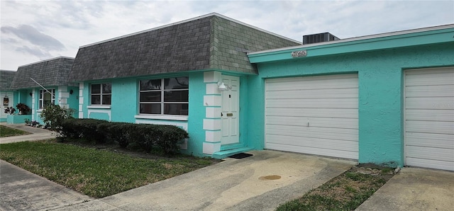 view of front of home with mansard roof, an attached garage, cooling unit, a shingled roof, and stucco siding