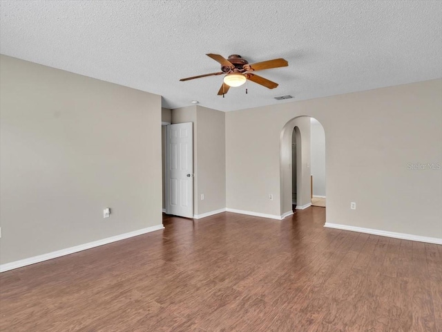 empty room featuring a ceiling fan, arched walkways, dark wood-style flooring, baseboards, and a textured ceiling