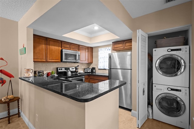 kitchen featuring a peninsula, stainless steel appliances, stacked washing maching and dryer, brown cabinets, and a raised ceiling