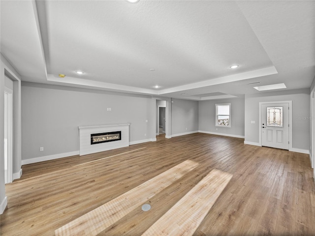 unfurnished living room featuring a tray ceiling, a glass covered fireplace, light wood-style flooring, and baseboards