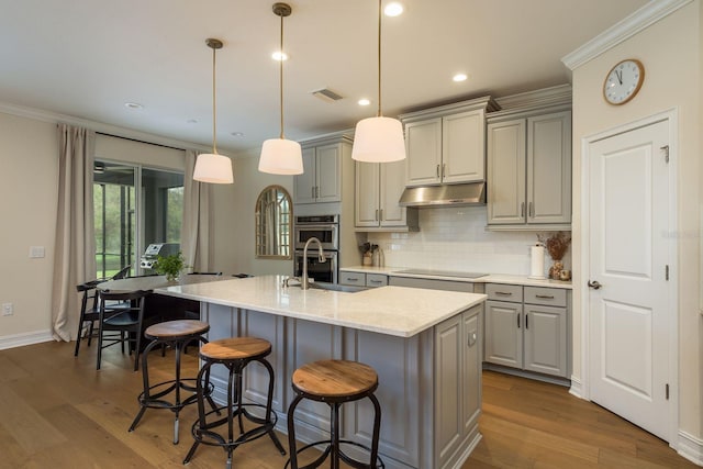 kitchen with a center island with sink, visible vents, light stone counters, hanging light fixtures, and under cabinet range hood