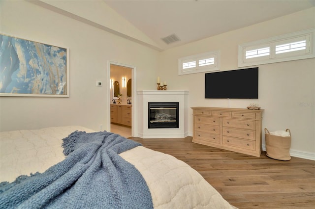 bedroom featuring lofted ceiling, connected bathroom, wood finished floors, visible vents, and a glass covered fireplace