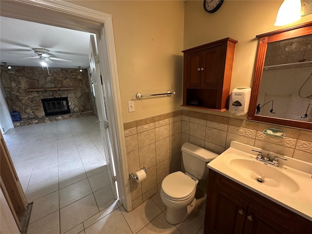 full bath featuring toilet, ceiling fan, a stone fireplace, tile patterned flooring, and vanity