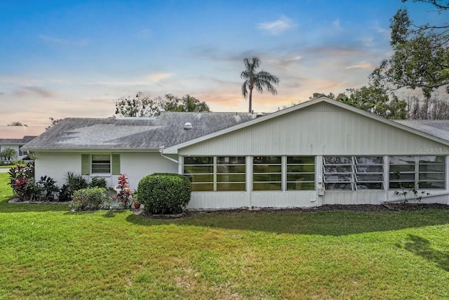 back of property at dusk featuring a sunroom, a shingled roof, stucco siding, and a yard