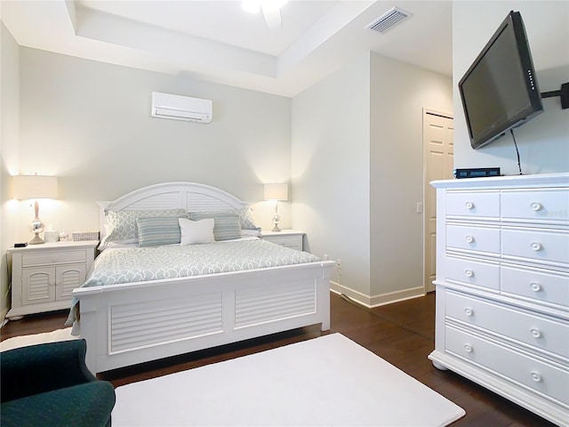 bedroom featuring dark wood-type flooring, a tray ceiling, a wall unit AC, and visible vents