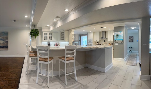 kitchen featuring visible vents, glass insert cabinets, crown molding, a kitchen breakfast bar, and wall chimney exhaust hood