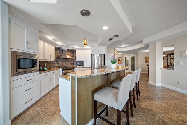 kitchen with a kitchen island, white cabinetry, appliances with stainless steel finishes, wall chimney exhaust hood, and a raised ceiling