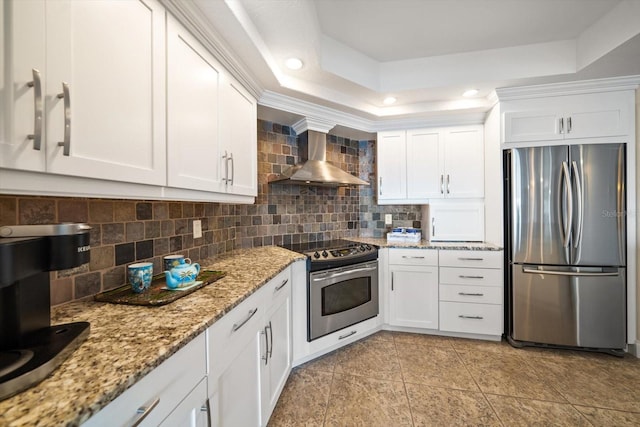 kitchen with appliances with stainless steel finishes, a raised ceiling, white cabinetry, and wall chimney range hood