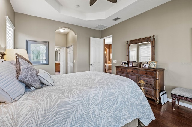 bedroom with dark wood-type flooring, a tray ceiling, multiple windows, and arched walkways