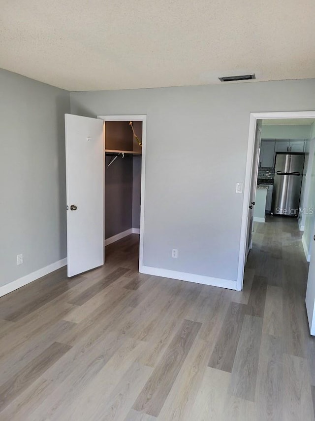 unfurnished bedroom featuring a textured ceiling, baseboards, freestanding refrigerator, light wood finished floors, and a walk in closet