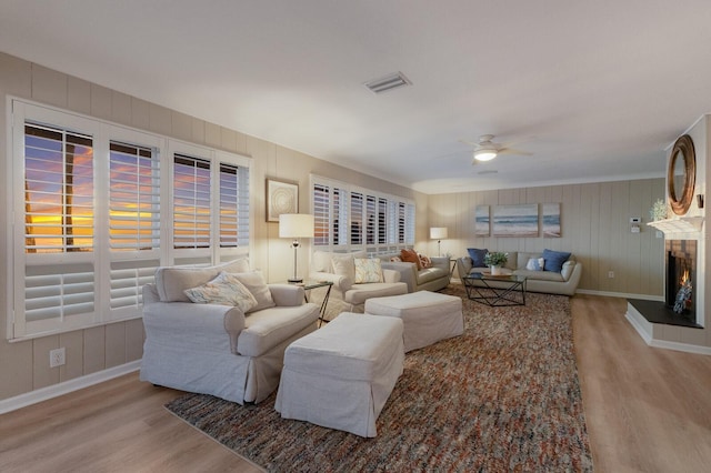 living room featuring light wood-style floors, a warm lit fireplace, visible vents, and ceiling fan