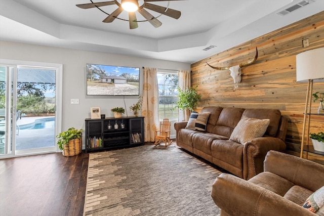 living room featuring a tray ceiling, dark wood-style flooring, visible vents, and wooden walls