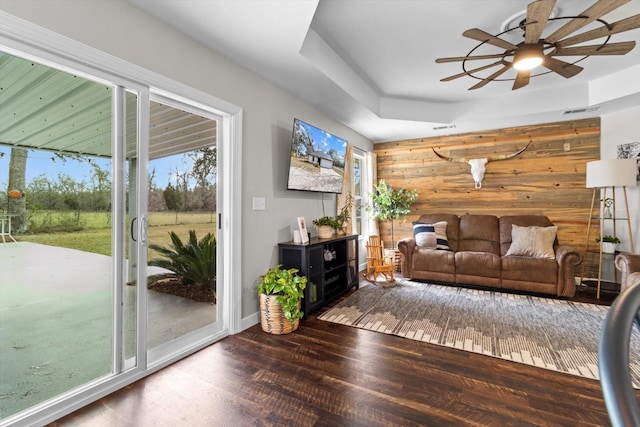 living room with wooden walls, visible vents, a raised ceiling, ceiling fan, and dark wood-type flooring