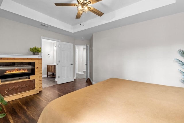 bedroom with dark wood-style flooring, visible vents, baseboards, a tray ceiling, and a glass covered fireplace