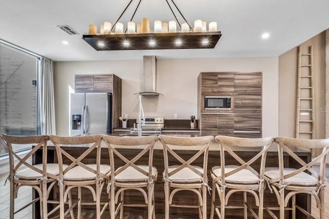 kitchen with wall chimney exhaust hood, modern cabinets, hanging light fixtures, built in microwave, and stainless steel refrigerator with ice dispenser