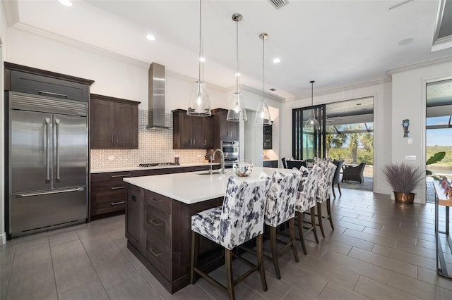 kitchen with a kitchen island with sink, stainless steel appliances, light countertops, wall chimney range hood, and pendant lighting