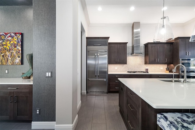 kitchen with dark brown cabinetry, wall chimney exhaust hood, appliances with stainless steel finishes, hanging light fixtures, and light countertops