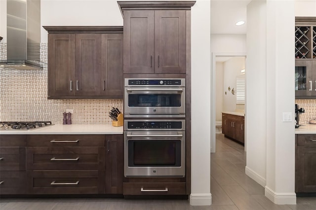 kitchen featuring tasteful backsplash, light countertops, appliances with stainless steel finishes, dark brown cabinetry, and wall chimney exhaust hood