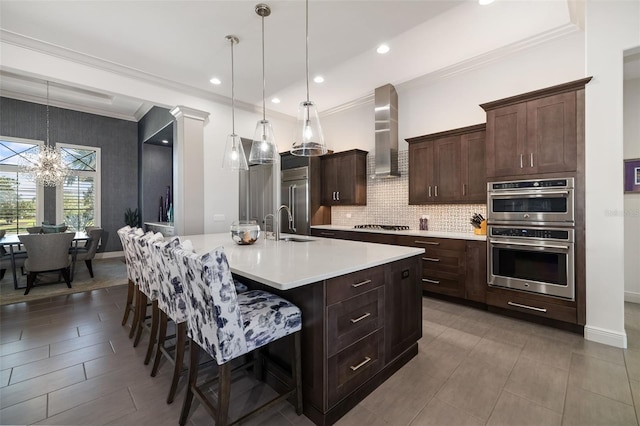 kitchen featuring light countertops, wall chimney range hood, appliances with stainless steel finishes, an island with sink, and a kitchen bar