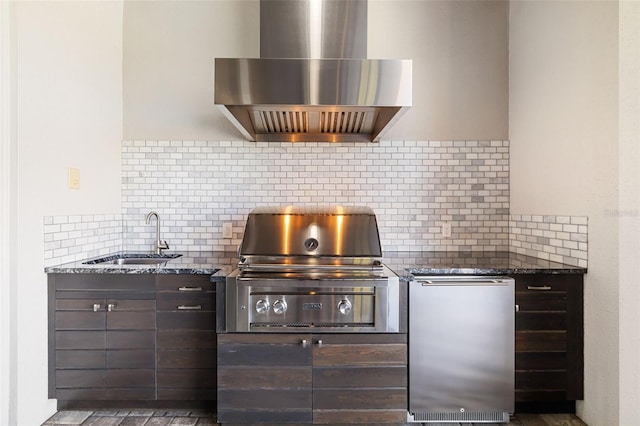 kitchen featuring a sink, dark brown cabinets, fridge, dark stone counters, and wall chimney exhaust hood