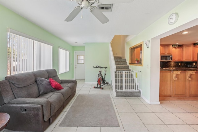 living room featuring light tile patterned flooring, visible vents, a ceiling fan, baseboards, and stairway