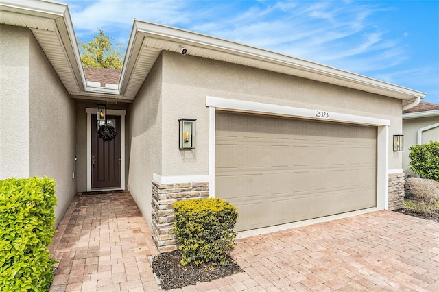 view of exterior entry featuring decorative driveway, an attached garage, stone siding, and stucco siding