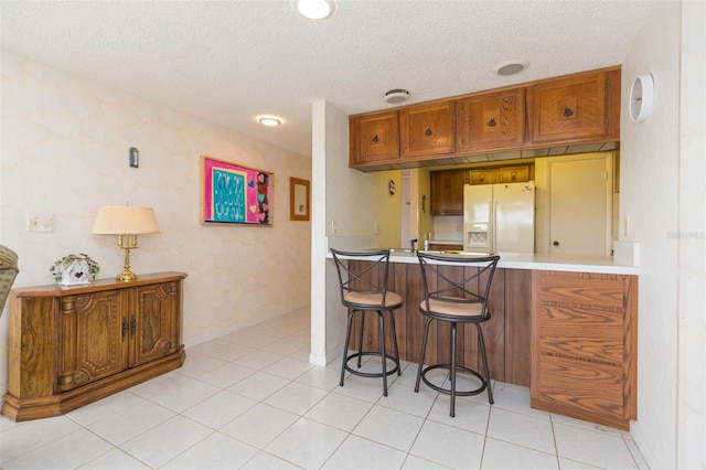 kitchen with white refrigerator with ice dispenser, brown cabinetry, a breakfast bar area, light countertops, and a textured ceiling