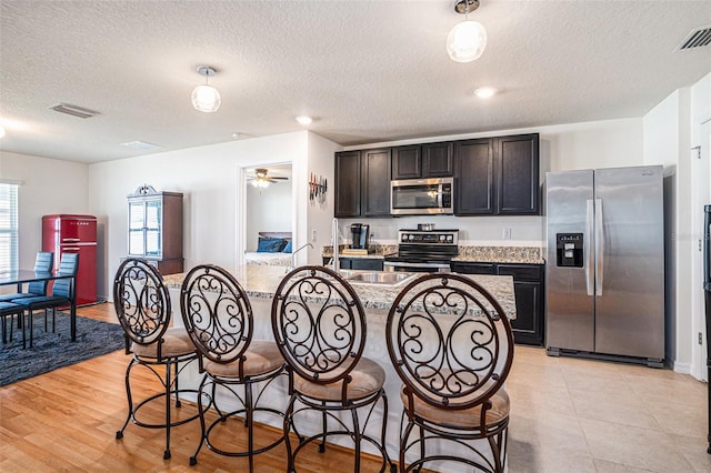 kitchen with a breakfast bar, visible vents, appliances with stainless steel finishes, a kitchen island with sink, and light stone countertops