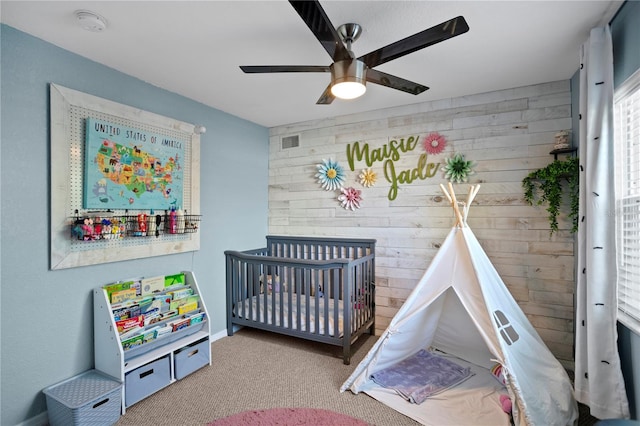 bedroom with wooden walls, visible vents, a ceiling fan, a nursery area, and carpet