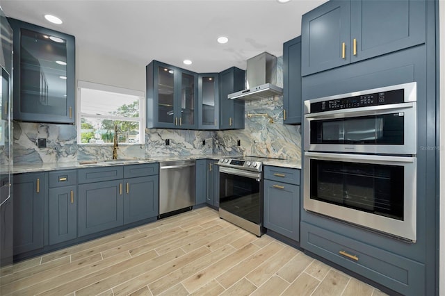 kitchen featuring tasteful backsplash, wall chimney exhaust hood, light wood-style flooring, appliances with stainless steel finishes, and a sink