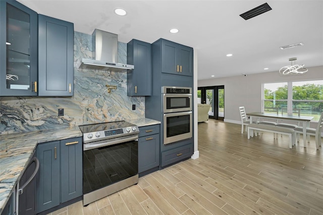 kitchen with light wood finished floors, decorative backsplash, appliances with stainless steel finishes, wall chimney range hood, and blue cabinets