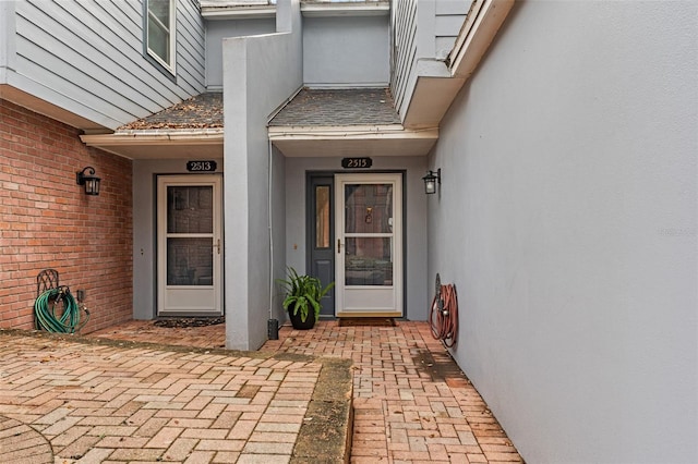 view of exterior entry with brick siding, roof with shingles, and stucco siding