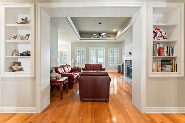 living area with light wood finished floors, a stone fireplace, a tray ceiling, and built in features