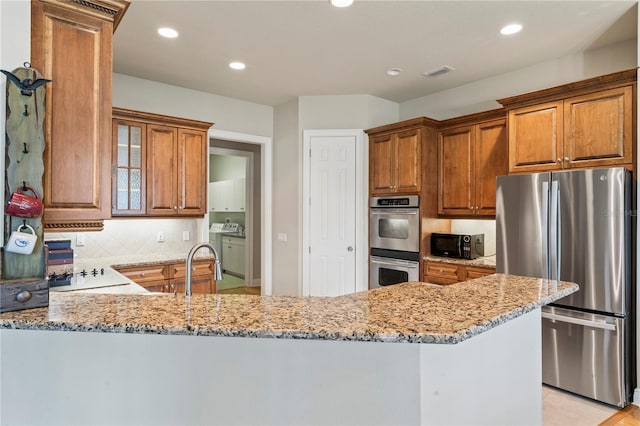 kitchen with light stone counters, a peninsula, appliances with stainless steel finishes, backsplash, and brown cabinets