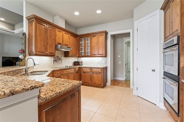 kitchen featuring brown cabinetry, glass insert cabinets, under cabinet range hood, double oven, and a sink