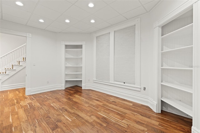 empty room with built in shelves, recessed lighting, stairway, wood finished floors, and baseboards