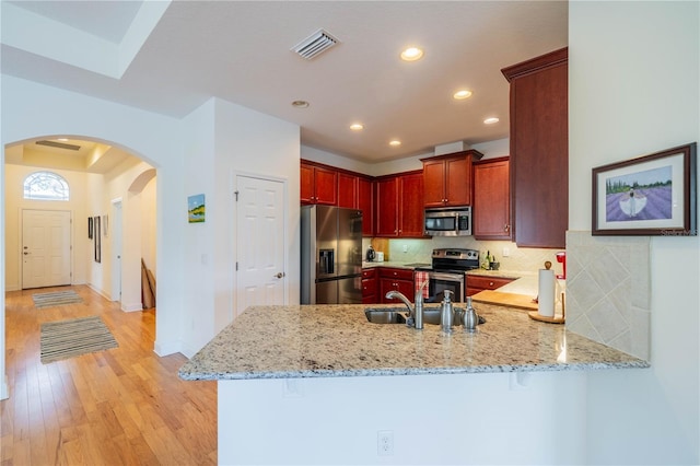 kitchen with reddish brown cabinets, stainless steel appliances, a sink, light stone countertops, and a peninsula