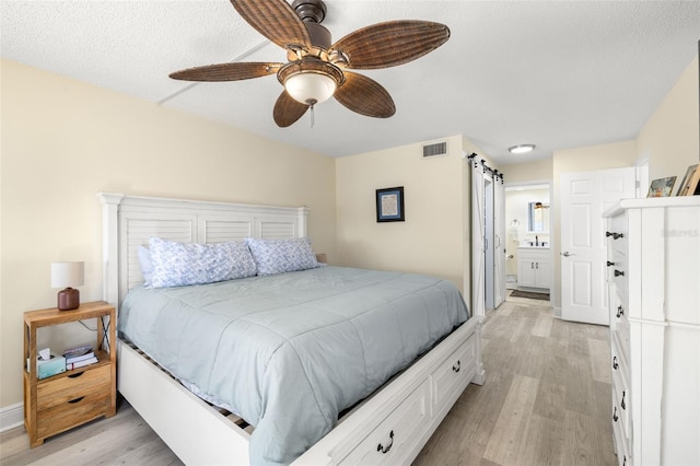 bedroom featuring ceiling fan, a barn door, visible vents, light wood-style floors, and ensuite bath