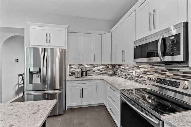 kitchen with arched walkways, white cabinets, light stone counters, appliances with stainless steel finishes, and backsplash