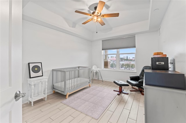 bedroom featuring a crib, baseboards, a raised ceiling, ceiling fan, and light wood-style flooring