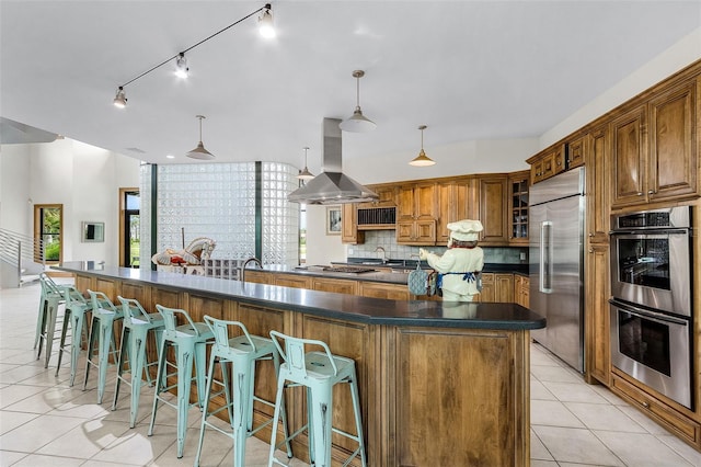 kitchen with island range hood, stainless steel appliances, brown cabinetry, dark countertops, and glass insert cabinets