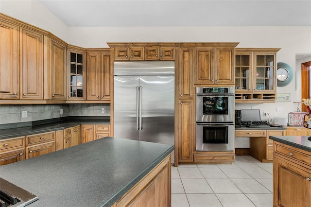 kitchen featuring light tile patterned floors, stainless steel appliances, dark countertops, glass insert cabinets, and brown cabinetry