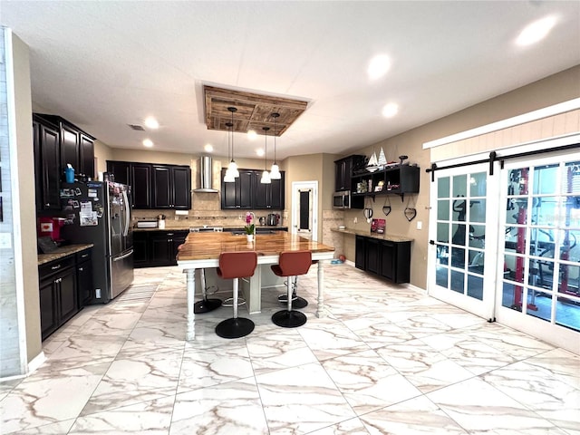 kitchen featuring wall chimney exhaust hood, wood counters, appliances with stainless steel finishes, dark cabinetry, and backsplash