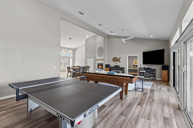 recreation room with a warm lit fireplace, baseboards, a ceiling fan, wood finished floors, and high vaulted ceiling