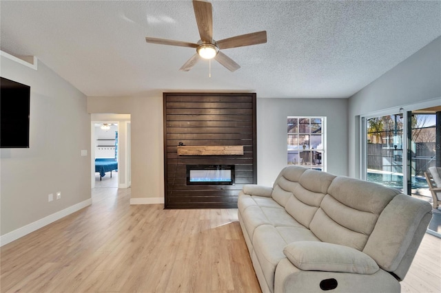 living room featuring ceiling fan, a textured ceiling, a large fireplace, baseboards, and light wood-type flooring