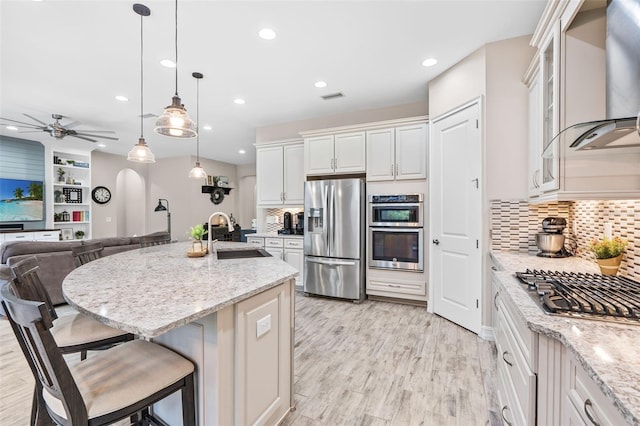 kitchen featuring light stone counters, stainless steel appliances, a sink, wall chimney exhaust hood, and light wood finished floors