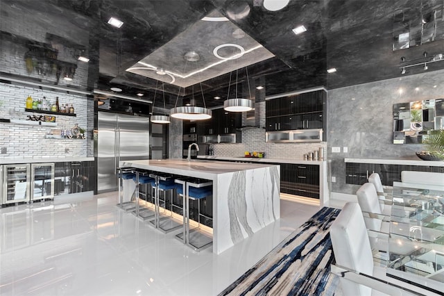 kitchen with an island with sink, appliances with stainless steel finishes, light countertops, dark cabinetry, and open shelves